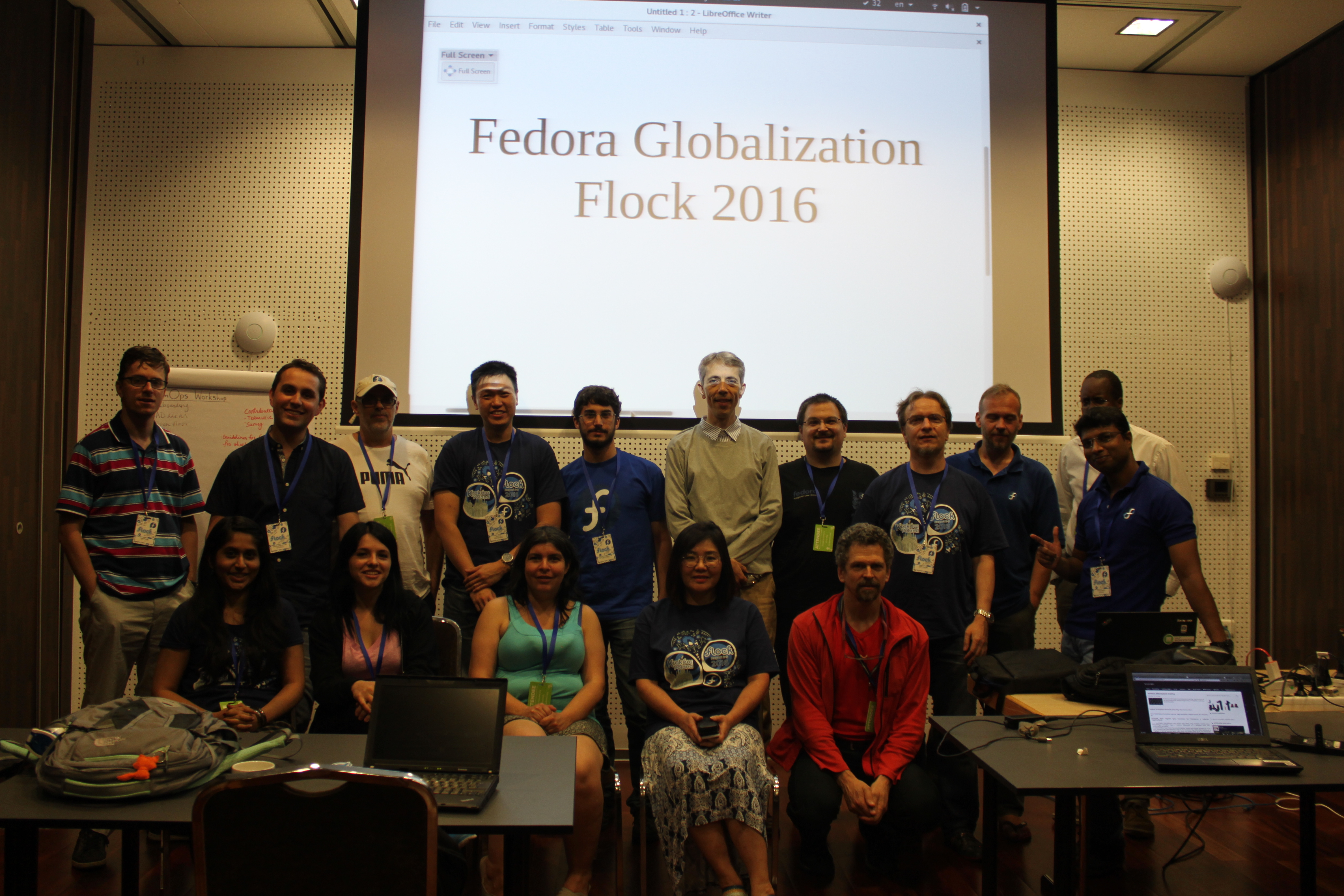 Группа из 15 человек, стоящих для фото перед экраном проектора со слайдом "fedora globalization flock 2016"