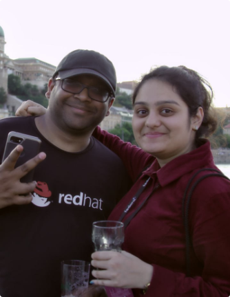 Deux personnes en extérieur souriant à la caméra. Le temps est nuageux. L'une des deux personnes porte une chemise noire avec le logo Red Hat Shadowman et faisant le signe "paix" avec sa main. L'autre porte un polo rouge et tient une boisson