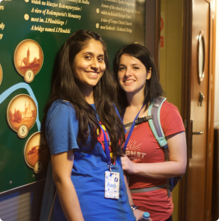 Deux personnes souriant debout devant un tableau d'information, elles portent chacune le badge de la conférence